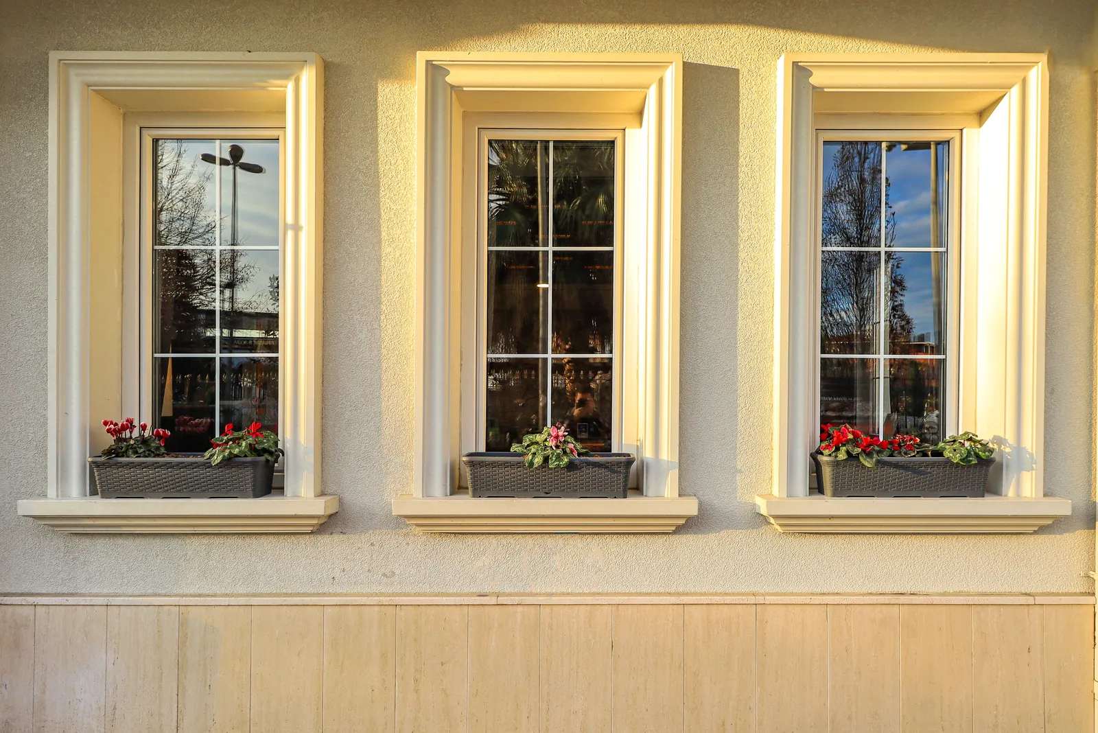 Three cream-framed exterior windows with white grille patterns and red flower boxes on a stucco wall at golden hour