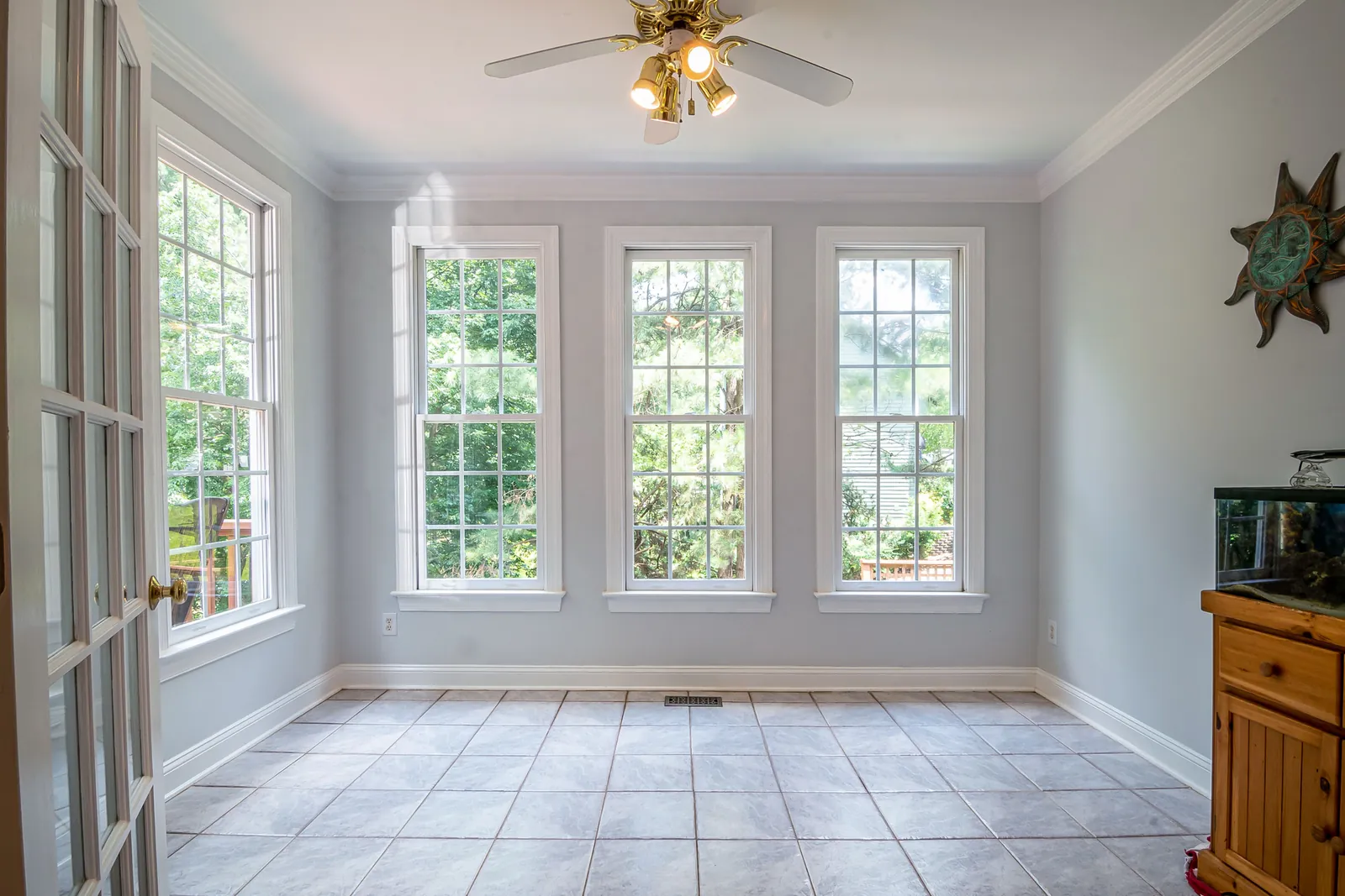 Sunlit residential room with three white double-hung windows and a glass-paned French door overlooking green trees